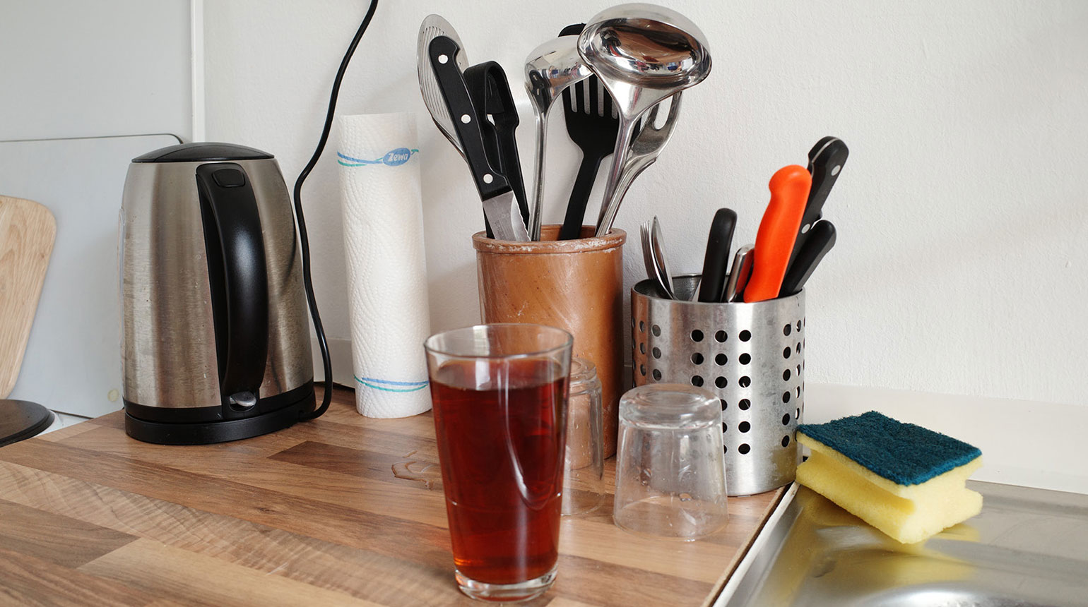 Nahaufnahme in einer Küche: ein Wasserkocher, Besteck in einem Behälter und ein Glas mit Tee.  