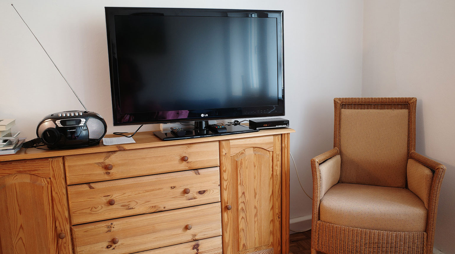 Detailaufnahme in einem Wohnraum: eine Kommode mit Fernseher und Radio, daneben ein Sessel. 
