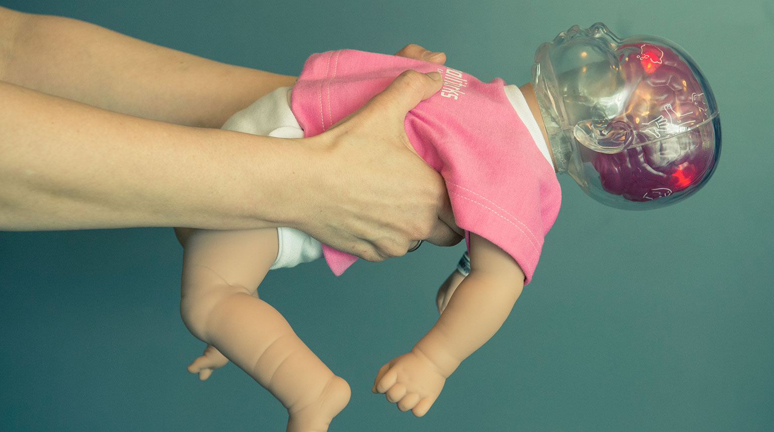 Nahaufnahme: Ein Mann hält eine „Shaken Baby“-Puppe. Der Körper wirkt echt, doch der Kopf ist aus durchsichtigem Plastik.