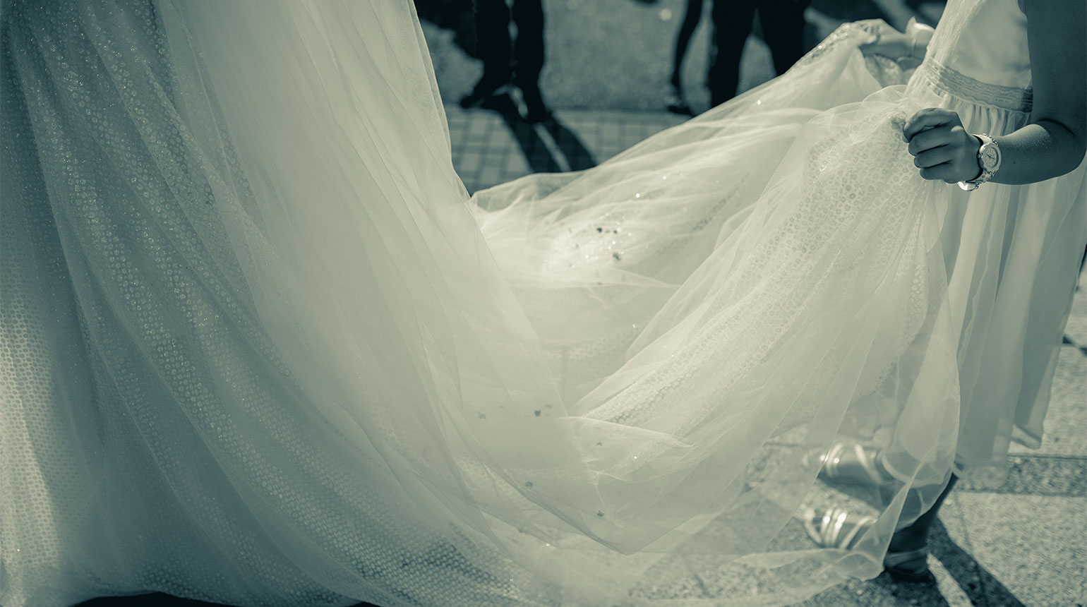 Szene auf einer Hochzeitsfeier. Zu sehen sind ein Teil des Brautkleids und die Beine der Umstehenden.