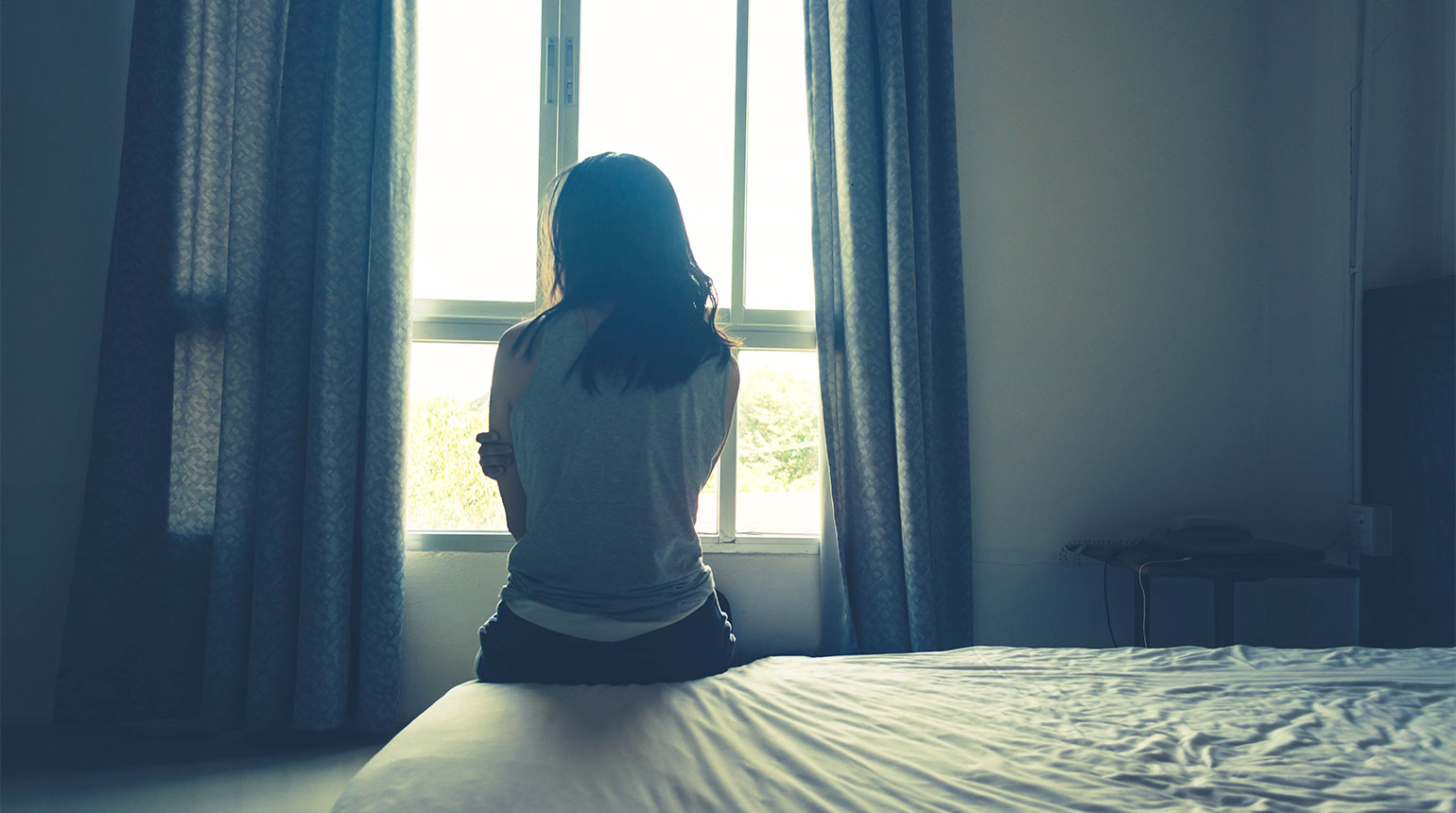 Eine junge Frau ist von hinten zu sehen. Sie sitzt auf einem Bett in einem düsteren Raum.