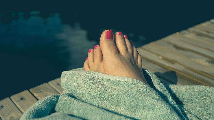 Auf einem Badesteg: Aus einem Badetuch gucken die nackten Füße einer Frau.