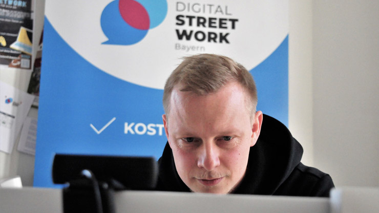 Nahaufnahme: Jonas Lutz schaut konzentriert auf seinen Bildschirm. Hinter ihm ist das Logo von Digital Streetwork Bayern. 