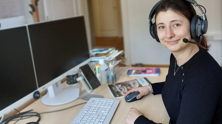 Porträtfoto: Jenny an einem Büro-Arbeitsplatz mit zwei Monitoren. Sie trägt ein Headset. 