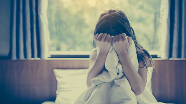 Eine Frau sitzt im Bett. Sie drückt die Bettdecke in einer verzweifelten Geste ans Gesicht.