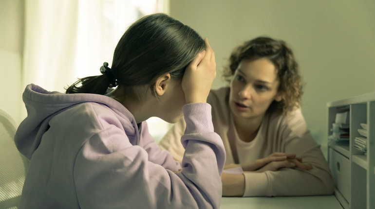 Eine Jugendliche stützt traurig den Kopf in die Hände. Ihre Mutter redet auf sie ein.