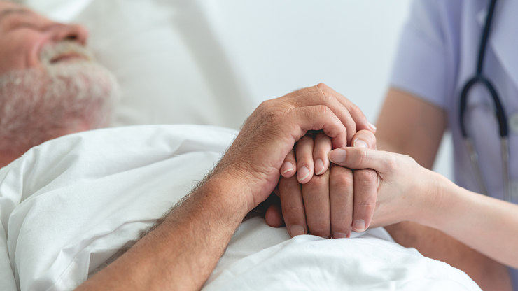 Eine Pflegekraft hält die Hände eines älteren, bettlägerigen Mannes.  