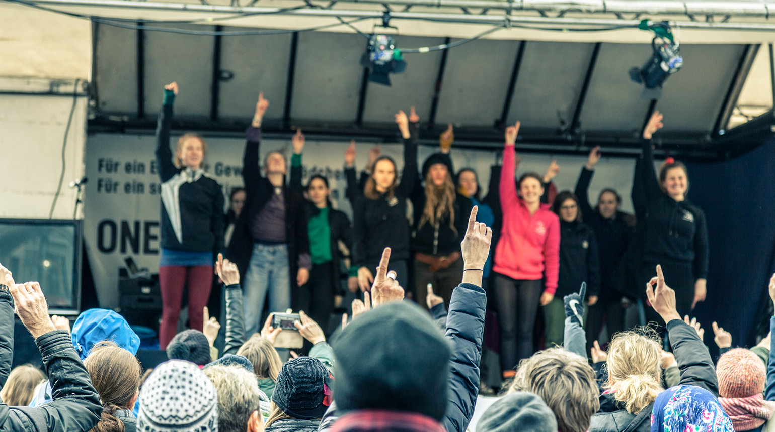 Im Freien: Mehrere Frauen stehen mit erhobenem Arm auf einer Bühne. Auch das Publikum hebt die Arme.