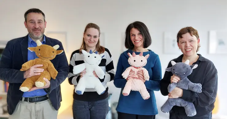 Gruppenbild: Vier Personen stehen nebeneinander. Sie halten freundliche „Monster“-Kuscheltiere im Arm.  