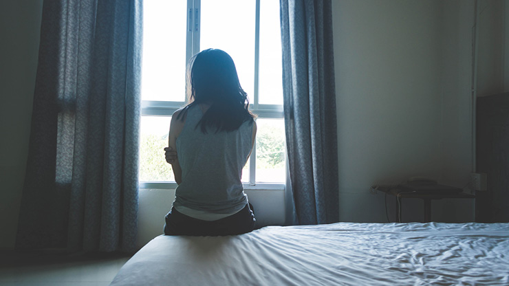 Eine junge Frau ist von hinten zu sehen. Sie sitzt auf einem Bett in einem düsteren Raum. 